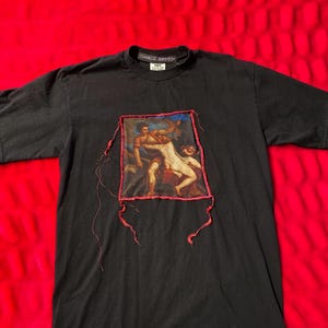 Pode incluir: T-shirt preto com um patch retangular com uma pintura em estilo renascentista. O patch é costurado na camisa com linha vermelha. A camisa tem o texto "CAMILLE SMOOCH" na gola.