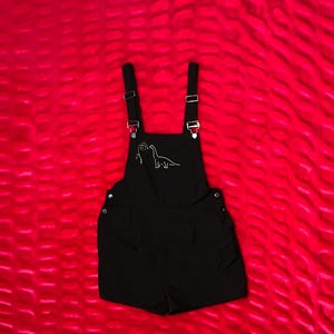 May include: Black overall shorts with adjustable straps and a white dinosaur outline on the bib. The shorts have two front pockets and are set against a red textured background.