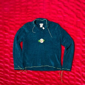 May include: Teal v-neck sweater with long sleeves. The sweater has a small snowman applique on the front and is trimmed with yellow stitching. The sweater is displayed on a red textured background.