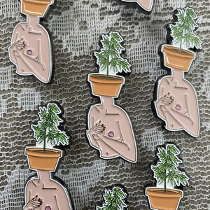 May include: Enamel pins featuring a nude figure with a potted plant for a head. The pins have a black outline, a tan body, and a pink nipple. The plant is green with a brown pot.