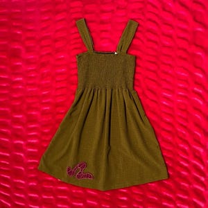 May include: Olive green dress with a smocked bodice and wide shoulder straps. The dress features a flared skirt and a magenta embroidered design near the hem. The fabric has a subtle textured pattern.
