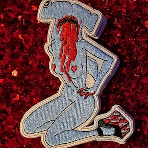 Peut inclure: Un patch brodé bleu représentant un requin marteau avec du sang rouge qui coule et un cœur sur la poitrine. Le requin est dans une pose séduisante, une jambe pliée et l'autre étendue. Le requin porte une chaussure à carreaux noirs et blancs.