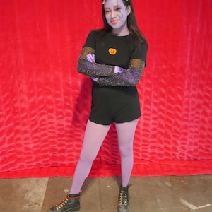 May include: A person in a Halloween costume stands in front of a red backdrop. They wear a black t-shirt with a pumpkin graphic, black shorts, and purple tights. The person's arms are crossed, and they wear black boots.