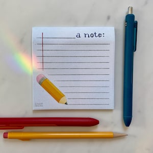 Puede incluir: Un bloc de notas blanco con líneas horizontales y el texto "a note:" se muestra con una ilustración de lápiz de dibujos animados. También hay un bolígrafo azul, un bolígrafo rojo y un lápiz amarillo. El bloc de notas tiene una línea vertical roja en el lado izquierdo.
