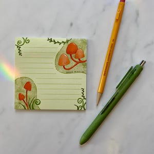 May include: A green and yellow notepad with a mushroom illustration and the text "Ginger Prints Co.". The notepad is on a white marble surface with a yellow pencil and a green pen.