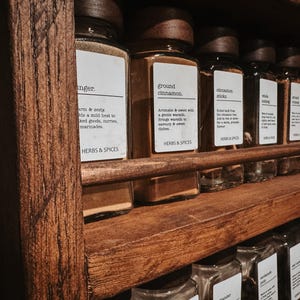 May include: A rustic wooden spice rack featuring multiple clear glass jars with dark wooden lids. Each jar has a white rectangular label with black text identifying the contents, such as ginger and ground cinnamon, and the brand Herbs & Spices.