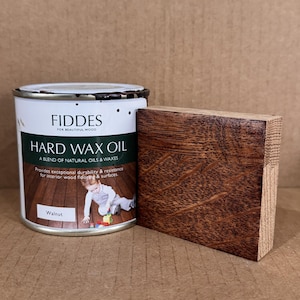 May include: A cylindrical metal can of Fiddes Hard Wax Oil in walnut finish, featuring a white label with green text and an image of a child on a wood floor. Next to the can is a rectangular block of wood stained in a dark walnut tone.