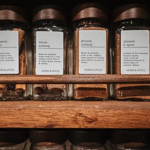May include: A row of clear glass spice jars with dark wooden lids, each featuring a white label with black text. The labels are marked with names including cinnamon sticks, whole nutmeg, ground nutmeg, and chinese 5 spice, all under the heading HERBS & SPICES.