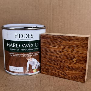May include: A white metal can of Fiddes Hard Wax Oil with a dark green label featuring text and a child on a wood floor, accompanied by a rectangular piece of wood stained in a medium brown tone with visible grain and a small knot.