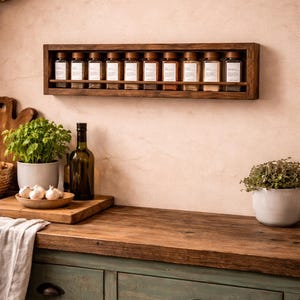 May include: A rectangular dark brown wooden wall-mounted spice rack holding ten clear glass jars with wooden lids. Each jar features a white rectangular label. The rack is mounted on a light-colored wall above a wooden countertop with green cabinets.