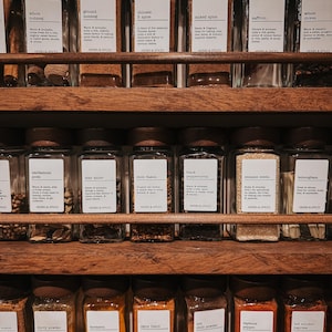 May include: A wooden spice rack holding three rows of clear glass jars with dark wooden lids. Each jar features a white label with black text identifying various herbs and spices like nutmeg, saffron, cloves, cardamom, and turmeric.