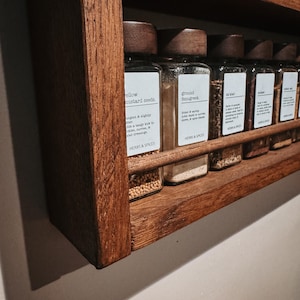 May include: A wall-mounted wooden spice rack with a dark brown finish, holding a row of clear glass spice jars with dark wooden lids. Each jar features a white rectangular label with black text identifying the contents and the brand Herbs & Spices.