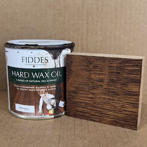 May include: A cylindrical metal can of Fiddes Hard Wax Oil in the color Whiskey, featuring a label with text and a child illustration, placed next to a rectangular wooden block stained in a dark brown tone with visible wood grain texture.