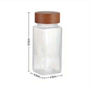 May include: A clear square glass jar with a brown wooden lid. The jar features technical dimension lines indicating a height of 10.5 cm and a width and depth of 4.5 cm, which is approximately 4.13 inches high and 1.77 inches wide and deep.