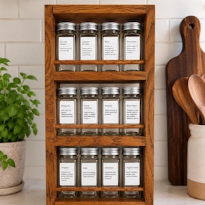 May include: A three-tiered wooden spice rack holding twelve clear glass jars with silver lids. Each jar features a white rectangular label with black text identifying spices such as Ground Ginger, Juniper Berries, Mace, Star Anise, Marjoram, Onion Granules, Nutmeg, Rose Petals, Oregano, Sage, Rosemary, and Nigella Seeds.