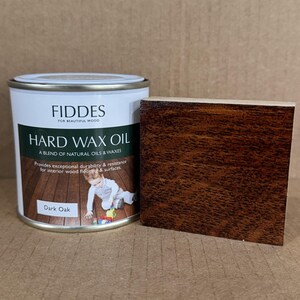 May include: A white metal can of Fiddes Hard Wax Oil in Dark Oak, featuring a label with text and a child playing on a wood floor, positioned next to a rectangular wood sample block stained in a matching dark oak finish.