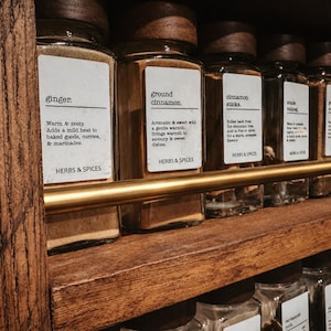 May include: A rustic wooden spice rack featuring clear glass jars with dark wooden lids. Each jar has a white label with black text identifying the contents like ginger and ground cinnamon, along with a brass-colored metal rail across the front.