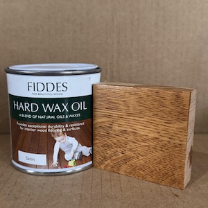 May include: A white metal can of Fiddes Hard Wax Oil with a dark green label featuring text and a child playing on a wood floor, accompanied by a rectangular wooden block with a smooth, light brown, satin-finish wood grain texture.