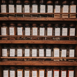 May include: A wooden spice rack with four shelves holding rows of clear glass jars with dark brown lids. Each jar features a white rectangular label with black text identifying various herbs and spices, organized in a neat, orderly display.