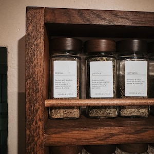 May include: A dark wood spice rack holding glass jars with dark wooden lids. The labels read thyme, parsley, and tarragon, each with descriptive text and the heading HERBS & SPICES. The rack features a horizontal wooden bar across the front.
