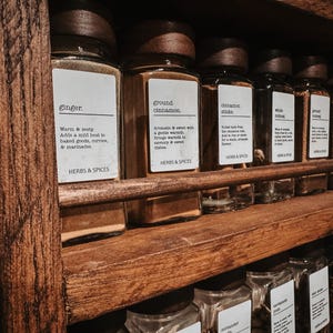 May include: A rustic wooden spice rack holding multiple clear glass jars with dark wooden lids. Each jar features a white rectangular label with black text identifying the contents, such as ginger and ground cinnamon, along with a brief description.