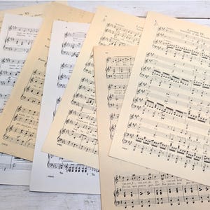 May include: A collection of vintage sheet music pages featuring black musical notation and English lyrics printed on aged, off-white paper. The individual sheets are layered and fanned out, displaying various piano lessons and musical exercises.