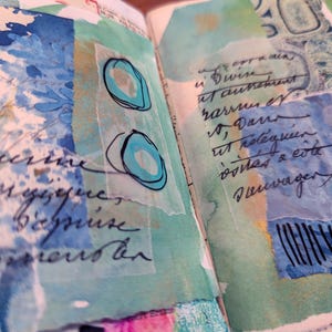 May include: An open journal page with mixed media art. The artwork features layered paper with watercolor washes in shades of blue and green. Handwritten script and abstract shapes are visible.