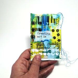 May include: A small, colorful, mixed media art journal page with the text "Creating art is" in black lettering on a yellow background.