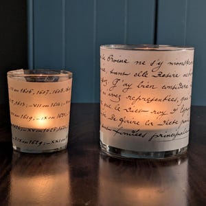 May include: Two glass candle holders with a vintage script design. The smaller holder is on the left, and the larger one is on the right. Both are lit, casting a warm glow. The text on the glass is in black.