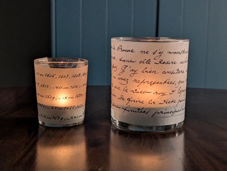 May include: Two glass candle holders with a vintage script design. The smaller holder is on the left, and the larger one is on the right. Both are lit, casting a warm glow. The text on the glass is in black.