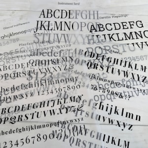May include: A collection of translucent sheets featuring black printed typography samples. The sheets display various font styles including Instrument Serif, Carollo Playscript, Glacial Indifference, Playfair Display, and Bauer Bodoni, showing uppercase and lowercase letters, numbers, and symbols.