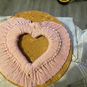 Może przedstawiać: R&oacute;żowa wisząca ozdoba ścienna w kształcie serca wykonana techniką makramy z frędzlami. Makrama jest zamontowana na okrągłym korkowym podkładzie.