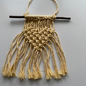 Może przedstawiać: Mały wiszący makramowy z sercem w środku. Wisiorek wykonany jest z naturalnego sznurka i przymocowany do małego drewnianego pręta.