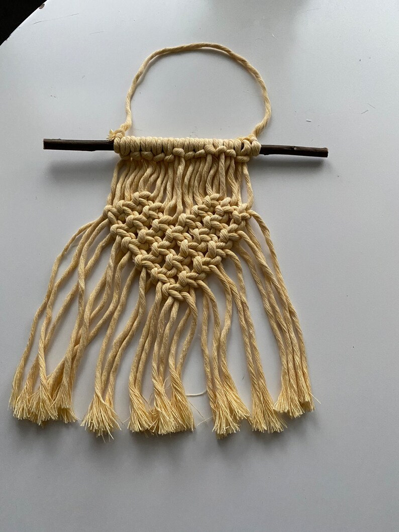 Może przedstawiać: Mały wiszący makramowy z sercem w środku. Wisiorek wykonany jest z naturalnego sznurka i przymocowany do małego drewnianego pręta.