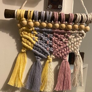 Może przedstawiać: Wisząca ozdoba ścienna wykonana techniką makramy z drewnianym drążkiem, ż&oacute;łtymi, fioletowymi i r&oacute;żowymi frędzlami oraz drewnianymi koralikami.