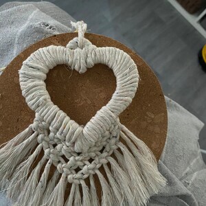 Może przedstawiać: Białe serce z makramu z frędzlami wiszące na korkowej podkładce. Serce jest wykonane z grubego białego sznurka z małymi kolorowymi plamkami.