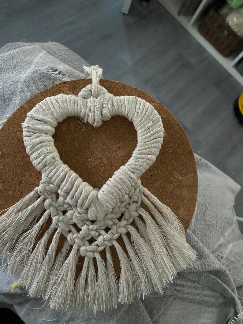 Może przedstawiać: Białe serce z makramu z frędzlami wiszące na korkowej podkładce. Serce jest wykonane z grubego białego sznurka z małymi kolorowymi plamkami.