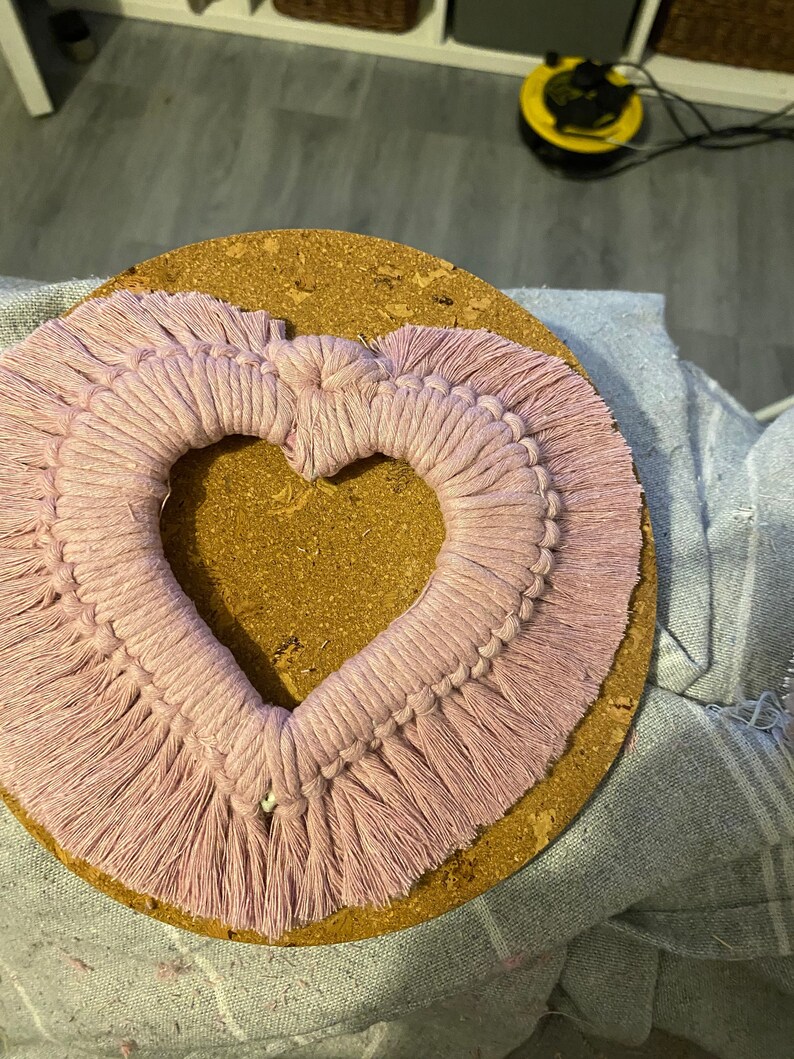 Może przedstawiać: R&oacute;żowa wisząca ozdoba ścienna w kształcie serca wykonana techniką makramy z frędzlami. Makrama jest zamontowana na okrągłym korkowym podkładzie.