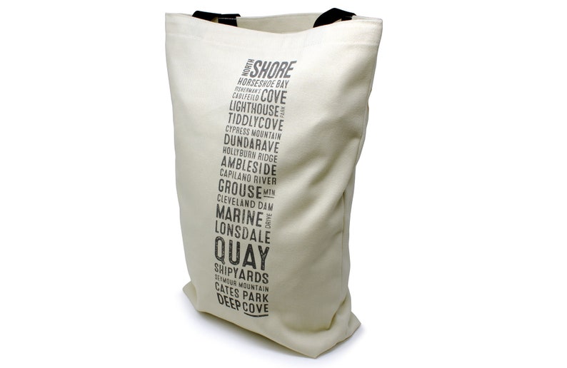 Puede incluir: Una bolsa de lona blanca con texto negro que enumera varios lugares en North Shore, Columbia Brit&aacute;nica. El texto dice "North Shore", "Horseshoe Bay", "Cholfield", "Lighthouse", "Tiddlycove", "Cypress Mountain", "Dundarave", "Hollyburn Ridge", "Ambleside", "Capilano River", "Grouse Min", "Cleveland Dam", "Marine Drive", "Lonsdale", "Quay", "Shipyards", "Seymour Mountain", "Cates Park" y "Deep Cove".