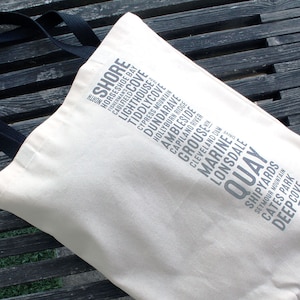 Puede incluir: Una bolsa de lona blanca con asas negras. La bolsa tiene un dise&ntilde;o de varios lugares impresos en ella, incluyendo "Shore", "Horseshoe Bay", "Caulfield Cove", "Lighthouse", "Tiddlycove", "Cypress Mountain", "Dundarave", "Hollyburn Ridge", "Ambleside", "Capilano River", "Grouse", "Cleveland Dam", "Marine", "Lonsdale", "Quay", "Shipyards", "Seymour Mountain", "Cates Park" y "Deep Cove".