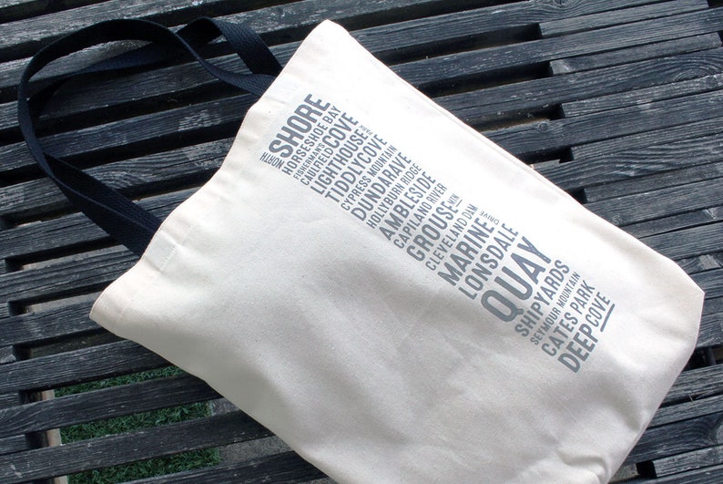 Puede incluir: Una bolsa de lona blanca con asas negras. La bolsa tiene un dise&ntilde;o de varios lugares impresos en ella, incluyendo "Shore", "Horseshoe Bay", "Caulfield Cove", "Lighthouse", "Tiddlycove", "Cypress Mountain", "Dundarave", "Hollyburn Ridge", "Ambleside", "Capilano River", "Grouse", "Cleveland Dam", "Marine", "Lonsdale", "Quay", "Shipyards", "Seymour Mountain", "Cates Park" y "Deep Cove".
