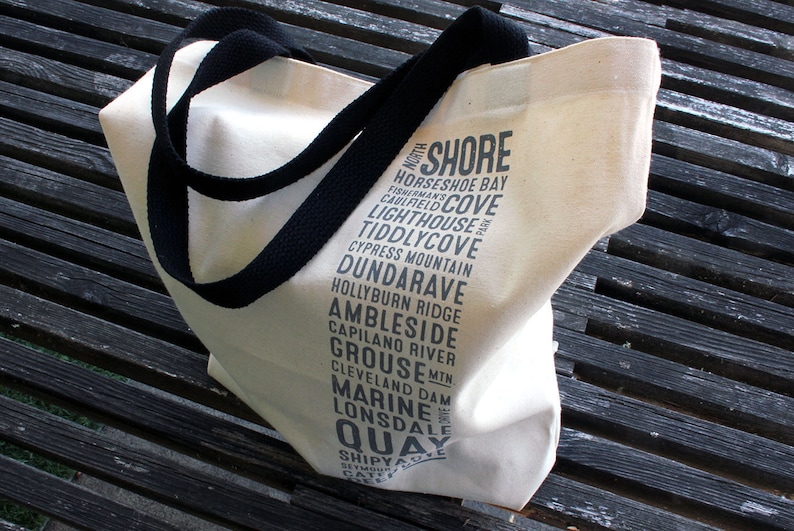 Puede incluir: Una bolsa de lona blanca con correas negras. La bolsa tiene texto negro impreso en ella, enumerando varios lugares en North Shore, Columbia Brit&aacute;nica, Canad&aacute;. El texto incluye: "SHORE", "HORSESHOE BAY", "CAULFIELD COVE", "LIGHTHOUSE", "TIDDLYCOVE", "CYPRESS MOUNTAIN", "DUNDARAVE", "HOLLYBURN RIDGE", "AMBLESIDE", "CAPILANO RIVER", "GROUSE", "CLEVELAND DAM", "MARINE", "LONSDALE", "QUAY", "SHIPYARD", "SEYMOUR", "CATHEDRAL".