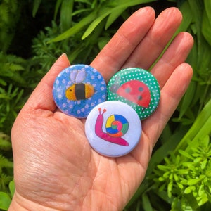 Puede incluir: Tres pines de botón coloridos con una abeja, un caracol y una mariquita. La abeja es amarilla y negra con alas marrones. El caracol es rosa con una concha de arcoíris. La mariquita es roja con manchas negras.