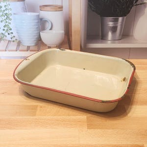 May include: A rectangular, off-white enamelware baking dish with a red trim. The vintage-style pan has rounded corners and is set on a wooden surface. The dish is empty and ready for use.