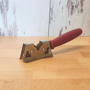 May include: Vintage metal and wood can sealer with a red wooden handle. The can sealer has a silver metal frame with two rollers and is designed to seal cans. The handle is a deep red color.