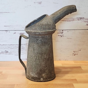 May include: An antique metal oil can with a spout and handle. The can is a weathered gray color, with the text "1 QT. LIQ." embossed on the side. The can is likely used for storing and pouring liquids.
