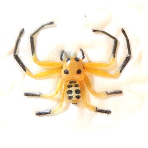 May include: A yellow spider figurine with black spots on its rounded body and head. The eight legs are yellow near the body and transition to black at the tips. The abdomen features a series of black horizontal stripes against a pale yellow base.