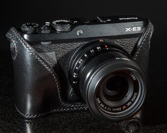 以下が含まれることがあります： 黒い革製のケースが付いた黒いカメラ。カメラには「SUPER EBC XF 23mm 1:2 R WR 043」と「HICAL LENS」の文字が印刷されたレンズが付いています。カメラの上部には「X-E3」の文字も印刷されています。