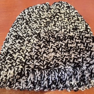 May include: A knitted beanie hat in a black and white speckled pattern. The hat features a folded brim and is made from a thick, textured yarn. The colour scheme is neutral, making it suitable for various outfits.