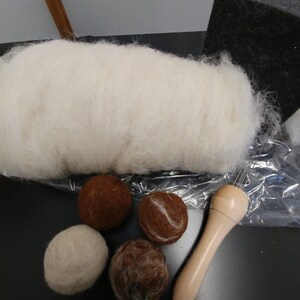May include: A collection of wool roving, felted wool balls in various shades of brown and white, a wooden needle tool, and a black foam block. The materials are for needle felting and crafting.